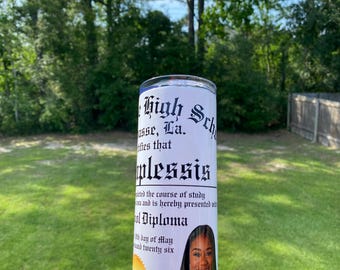 May include: A white tumbler with a graduation theme. The tumbler features text in a gothic font, a photo of a smiling person, and a gold seal. The text includes the words "High School Diploma".