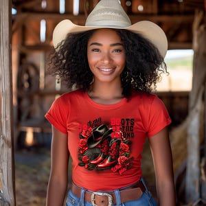 May include: A red t-shirt with a graphic of red cowboy boots, a black cowboy hat, and red roses. The text on the shirt reads "I got my boots on the ground, who's got them feet up?"