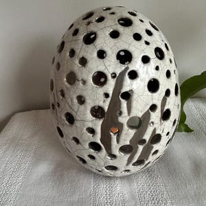 Puede incluir: Una esfera de cerámica blanca con un acabado desgastado y muchos agujeros pequeños. La esfera tiene un esmalte marrón en el centro y es un objeto decorativo.