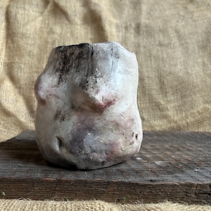 May include: A handmade, organic-shaped ceramic vase. The vase has a rough, textured surface with a mix of white, gray, and hints of purple and brown. The top edge is dark, and the vase sits on a wooden surface.