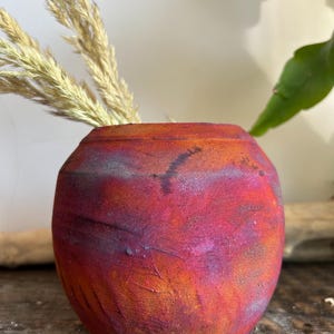 May include: A small, round ceramic vase with a textured surface. The vase is painted with a blend of red, orange, and purple hues. Dried grasses are arranged inside the vase, adding a natural element to the composition. The vase is approximately 4 inches tall.