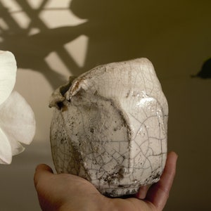 Puede incluir: Una maceta de cerámica blanca con un esmalte craquelado, sostenida en una mano. La maceta tiene una forma irregular y una textura rugosa.