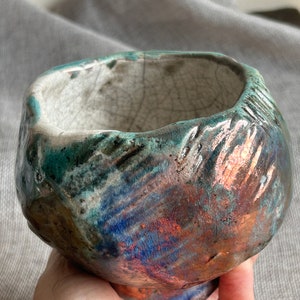 Peut inclure: Un petit bol rond en céramique avec un glaçage texturé et craquelé dans des tons de bleu, vert et cuivre. Le bol a une forme unique et irrégulière et une base légèrement surélevée.