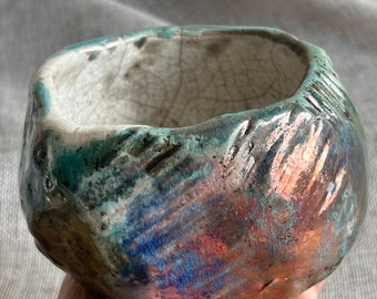 May include: A small, round ceramic bowl with a textured, crackled glaze in shades of blue, green, and copper. The bowl has a unique, irregular shape and a slightly raised base.