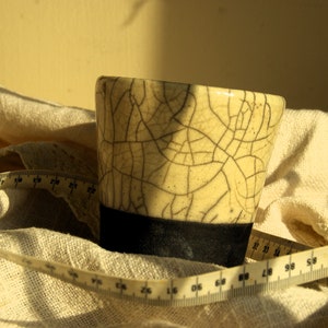 Puede incluir: Una taza de cerámica blanca con un esmalte craquelado y un borde negro. La taza está sentada sobre una tela blanca con una cinta métrica.