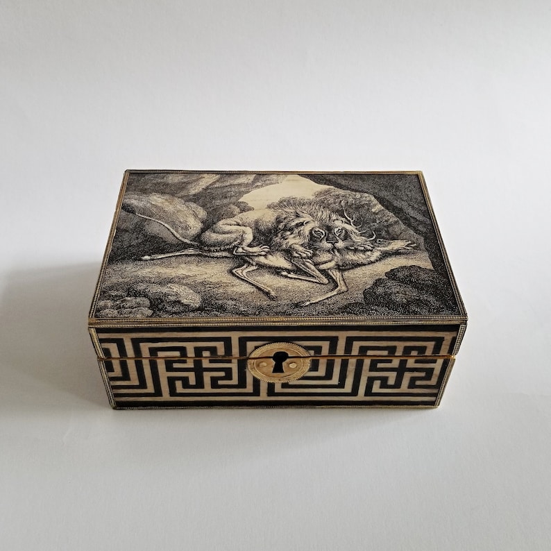 May include: A rectangular box with a detailed black and white illustration on the lid, depicting a hunting scene. The box features a geometric pattern around the base and a gold-colored keyhole.