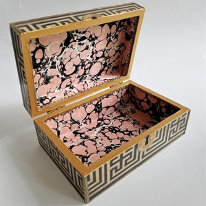 May include: Open rectangular box with a hinged lid. The exterior features a black and white geometric pattern, while the interior is lined with pink, black, and white marbled paper. The box has gold-colored trim and a small clasp.