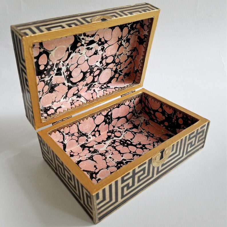 May include: Open rectangular box with a hinged lid. The exterior features a black and white geometric pattern, while the interior is lined with pink, black, and white marbled paper. The box has gold-colored trim and a small clasp.
