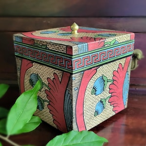 May include: A square decorative box with a gold finial on the lid. The box features a repeating Greek key pattern in green and blue, with red floral and leaf designs. The box is made of wood and has a vintage aesthetic.