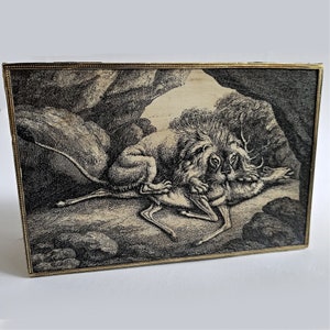 May include: A framed black and white etching depicts a lion attacking a deer in a cave setting. The artwork has a detailed, vintage style, with a gold-colored frame. The scene is rendered with fine lines and shading, creating a dramatic effect.
