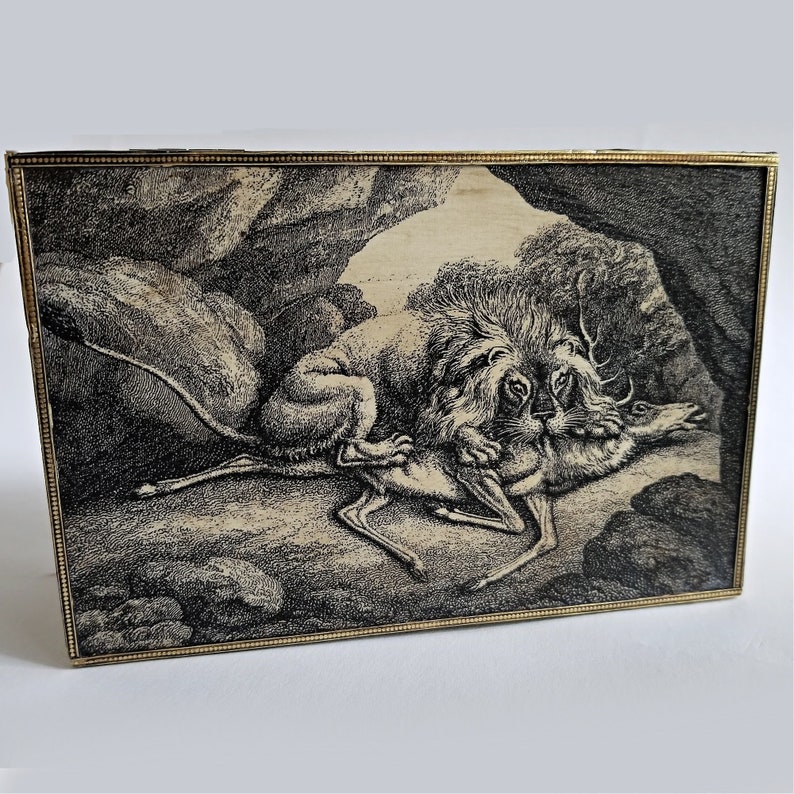May include: A framed black and white etching depicts a lion attacking a deer in a cave setting. The artwork has a detailed, vintage style, with a gold-colored frame. The scene is rendered with fine lines and shading, creating a dramatic effect.