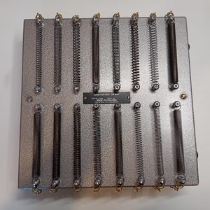 May include: A rectangular grey metal junction box featuring sixteen vertical metal tension springs arranged in two rows. A central black rectangular plate displays the text: JUNCTION BOX 48 WAY, TYPE No. 2750A, SERIAL No., MADE IN ENGLAND, THE M-O VALVE LTD. HAMMERSMITH.