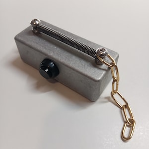 Può includere: Una scatola di metallo color argento con un pomello nero e una molla fissata con viti. È attaccata una catena color oro. L'oggetto è su una superficie bianca.