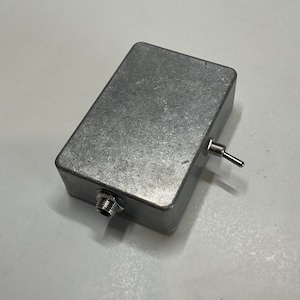 Può includere: Un contenitore rettangolare in alluminio spazzolato dotato di un interruttore a levetta argentato sul lato e di una porta jack di ingresso circolare filettata sul bordo anteriore. La superficie presenta una finitura industriale grezza.