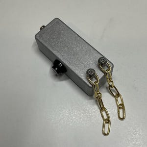 May include: A rectangular silver-coloured metal enclosure featuring a single black circular knob on the side and two gold-toned metal chains attached to the top surface by two silver-coloured bolts. The device has a smooth, matte finish.