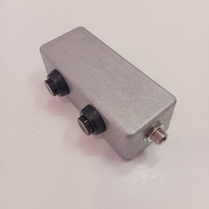 May include: A rectangular, silver electronic device featuring two black knobs and a threaded connector. This component, likely for audio or electronics, has a metallic finish and a compact design.