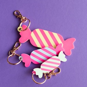 May include: Three candy-shaped keychains with pink bodies and striped designs in yellow, blue, and white. Each keychain has a gold-colored clasp and ring. The background is a solid purple.