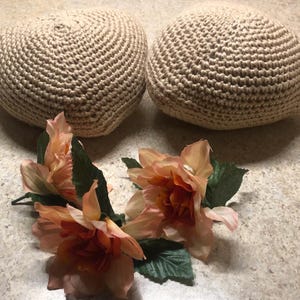May include: Two beige, crocheted, round objects are displayed with a cluster of peach-colored artificial flowers with green leaves. The crocheted items have a textured surface. The flowers are in the foreground.