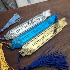 Może przedstawiać: Trzy dekoracyjne zakładki do książek z chwostami. Jedna jest srebrna z hebrajskim tekstem, druga niebieska z "EMILY | MAZEL TOV!", a trzecia złota z "JACOB | MAZEL TOV!". Każda zakładka ma chwost w innym kolorze.