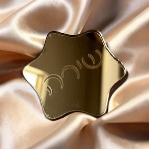 May include: A gold-colored, star-shaped mirror with etched Hebrew text. The mirror has a reflective surface and is set against a backdrop of a satin fabric.