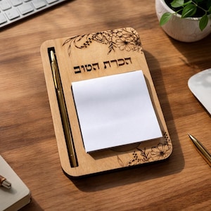 May include: A rectangular light wood notepad holder featuring a gold pen in a side slot and a stack of white sticky notes. The wood surface is engraved with floral patterns at the top and bottom corners and the Hebrew text "הכרת הטוב".