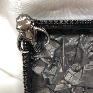 May include: A black picture frame with a silver metal embellishment in the shape of a creature with tusks. The frame is decorated with black rhinestones and the inside is lined with black tissue paper with iridescent glitter.
