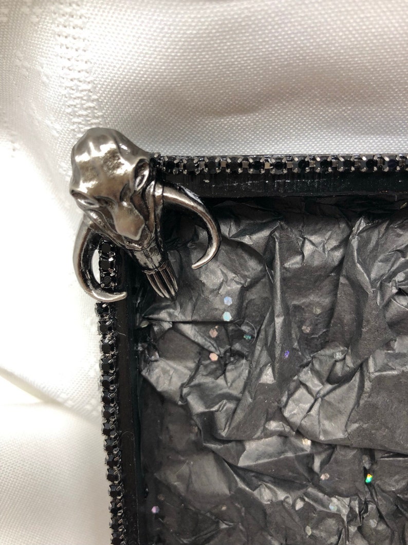 May include: A black picture frame with a silver metal embellishment in the shape of a creature with tusks. The frame is decorated with black rhinestones and the inside is lined with black tissue paper with iridescent glitter.