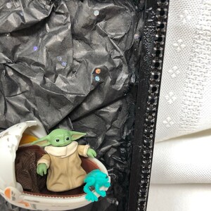 May include: A small green figure in a brown robe sits in a white egg-shaped pod with a blue frog figurine next to it. The pod is on a clear stand against a black background with silver glitter.