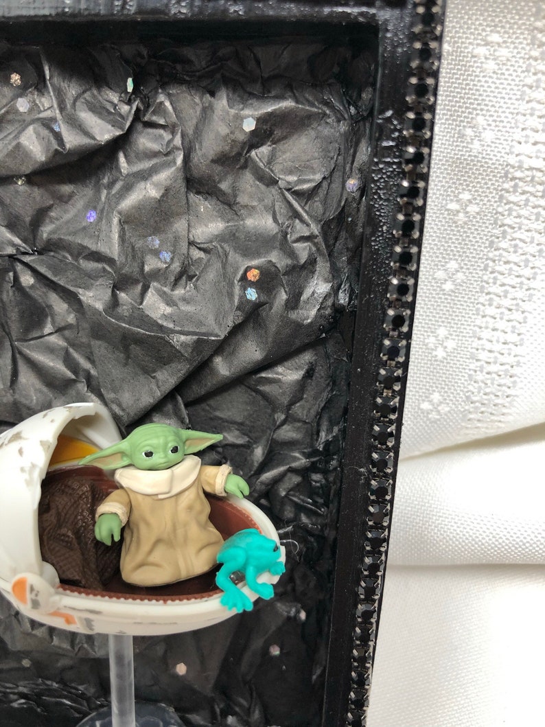 May include: A small green figure in a brown robe sits in a white egg-shaped pod with a blue frog figurine next to it. The pod is on a clear stand against a black background with silver glitter.