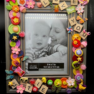 May include: A gray picture frame decorated with colorful buttons, candies, and other embellishments. The frame is 5 inches by 7 inches and has the words "LOVE YOU Cutie" spelled out with letter tiles. A photo of a mother and child is inside the frame.