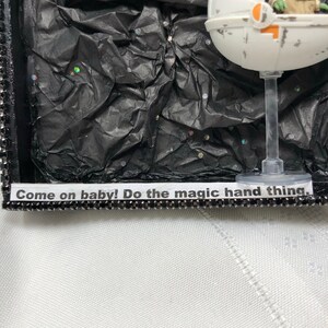 May include: A small, green, fictional character sits in a white, round, spaceship-like pod. The pod is on a clear stand against a black background with silver glitter. The text "Come on baby! Do the magic hand thing." is on a black and silver strip below the pod.