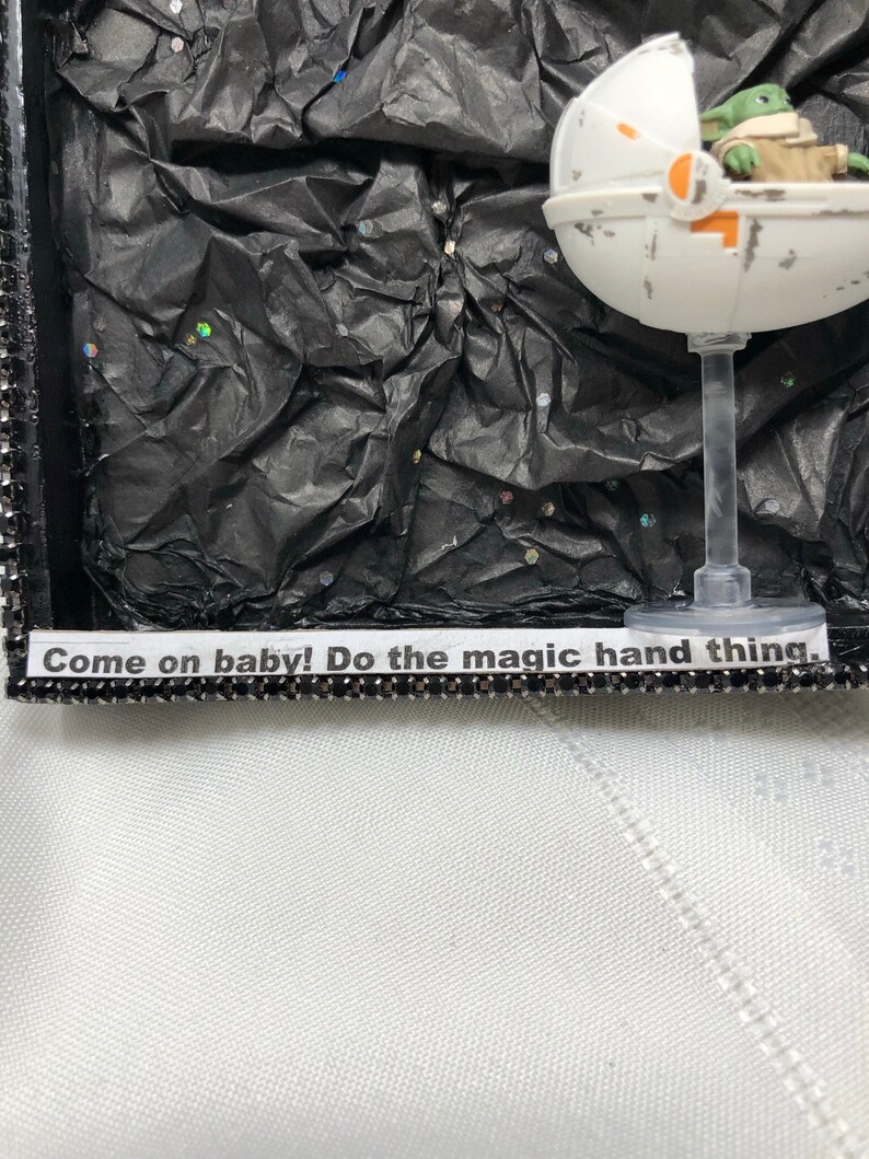 May include: A small, green, fictional character sits in a white, round, spaceship-like pod. The pod is on a clear stand against a black background with silver glitter. The text "Come on baby! Do the magic hand thing." is on a black and silver strip below the pod.