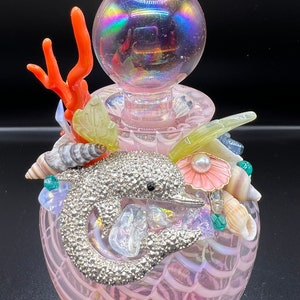 May include: A pink glass bottle with a iridescent stopper and a dolphin charm. The bottle is decorated with seashells, coral, and other ocean-themed embellishments.