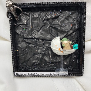 May include: A framed shadow box with a black background and a small green alien figure in a clear plastic pod. The pod is on a clear plastic stand. The alien figure is holding a small green object. The shadow box has a black frame with a silver trim. The text "Come on baby! Do the magic hand" is on a white label at the bottom of the shadow box.
