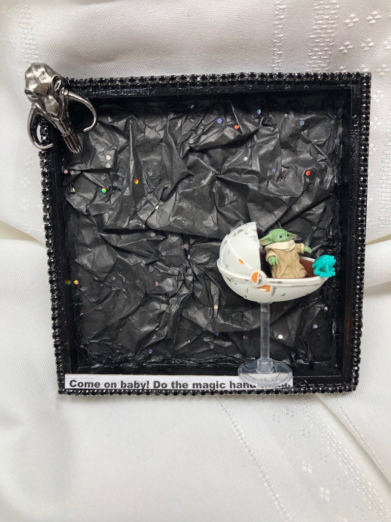 May include: A framed shadow box with a black background and a small green alien figure in a clear plastic pod. The pod is on a clear plastic stand. The alien figure is holding a small green object. The shadow box has a black frame with a silver trim. The text "Come on baby! Do the magic hand" is on a white label at the bottom of the shadow box.