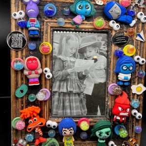 May include: A wooden picture frame decorated with colorful characters and objects from the movie Inside Out. The frame features a black and white photo of two children looking at a piece of paper. The frame is decorated with various characters, including Joy, Sadness, Anger, Fear, and Disgust. The frame also features a variety of other objects, including googly eyes, glitter, and small toys.