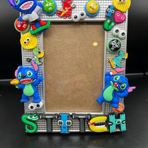 May include: A silver picture frame decorated with colorful 3D characters and embellishments. The frame features the word "STITCH" spelled out with colorful letters and includes multiple images of the character Stitch from the Disney movie Lilo & Stitch.