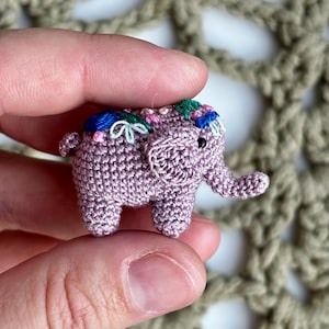 May include: A small, crocheted elephant in a muted purple colour. It features embroidered details in blue, green, and pink on its back and head. The elephant has a trunk, ears, and four legs. The craftwork is intricate and detailed.
