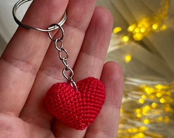 May include: A red crocheted heart keychain with a silver-colored chain and keyring. The heart is a vibrant red color and appears to be handmade. The keychain is held in a person's hand.
