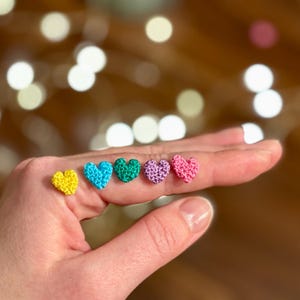 May include: A hand displaying six small, crocheted hearts in various colors: yellow, turquoise, green, purple, and pink. The hearts are evenly spaced across the palm and fingers, with a blurred background of soft, out-of-focus lights.