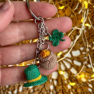 Peut inclure: Un porte-clés sur le thème de la Saint-Patrick avec un chapeau haut de forme vert crocheté avec une bande jaune, un pot d'or marron et un trèfle vert. Le porte-clés est attaché à un anneau et une chaîne argentés.