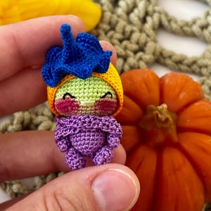 May include: A handmade crochet doll with a smiling face, wearing a blue flower hat, yellow cap, and purple outfit. The doll is held in a hand, with an orange pumpkin in the background. The doll is approximately 2 inches tall.
