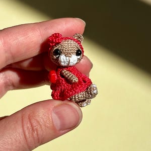 Puede incluir: Una figura de oso en miniatura tejida a ganchillo con hilo marrón claro, hocico blanco y ojos de cuentas negras. El oso lleva un vestido rojo brillante tejido a ganchillo, una flor roja a juego en la cabeza y un pequeño corazón rojo al costado.