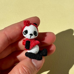 Può includere: Una piccola figura di orsetto panda all'uncinetto. Il panda ha una testa bianca con dettagli neri intorno agli occhi e alle orecchie, e una fascia rossa. Indossa una giacca rossa e ha gambe e piedi neri. Articolo fatto a mano.
