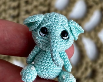 May include: A small, crocheted elephant in a light blue color. The elephant has large, black eyes with white highlights, and white accents on its feet. The handmade toy is a miniature version of an elephant.