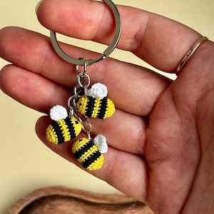 May include: A metal keychain featuring three small, crocheted bee charms. Each bee is crafted with alternating horizontal stripes of yellow and black yarn, and each features a pair of small, white crocheted wings attached to the top of the body.