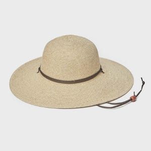 May include: A beige sun hat with a wide brim and a rounded crown. The hat features a brown leather band around the crown and a chin strap with a wooden bead. The hat is made of woven material.