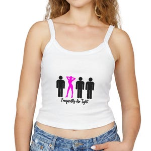 Op de afbeelding: Witte tanktop met een afbeelding van drie stokfiguren, waarvan één roze, en de tekst "Frequently Are Tight". De top heeft dunne bandjes en een ronde halslijn. Het model draagt een blauwe spijkerbroek.