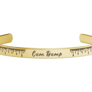 Könnte beinhalten: Ein goldfarbenes Armreif-Armband mit dem Text "Cum Dump" in handschriftlichem Stil eingraviert. Das Armband hat ein tropfendes Design entlang der Kanten.