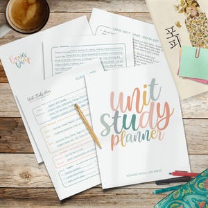 May include: A collection of educational materials, including a "Unit Study Planner" with colorful text, and other worksheets. A gold pen rests on the planner. The items are arranged on a wooden surface, with a cup of coffee in the upper left corner.